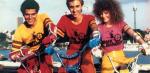 Three teenagers in brightly coloured clothes sitting on BMX bikes and smiling at the camera.