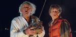 An older man with white hair, wearing a lab coat and holding a large remote control standing next to a young man at night time, they both look shocked as they stare into the distance.