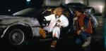 An older man with white hair, wearing white cover-alls and sitting in a DeLorean car while a young man kneels on the ground next to the car talking to him.