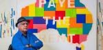 An older First Nations man pictured seated in front of a map of Australia on the wall. The words 'Paye the Rent' are painted over the map.
