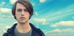 Head and shoulders of a young man looking intently into the camera against a blue sky dotted with clouds.