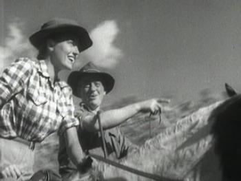 Film still of a woman and a man on horseback. The man points into the distance