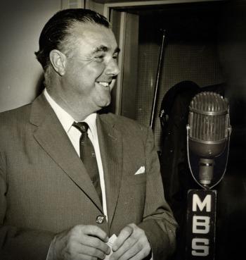Radio announcer Terry Dear, circa 1960, standing next to a microphone with the MBS logo on it. He is looking sideways and smiling.