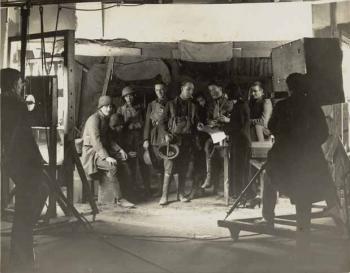 A group of actors stand on a set. There is a camera man in the foreground with a large old-fashioned camera. A woman is directing the action. You can only just make out her silloughette because she is in a long black dress with a short black bob.
