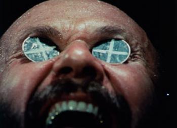 Close up of a mans face, mouth open, with two-up coins with crosses on them in his eye sockets