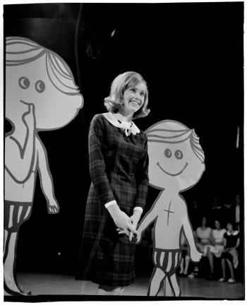 Olivia Newton John on GO!! Singer Olivia Newton-John on stage in a TV studio in an episode of the Go Show. She is pictured full length, standing between two lifesize cartoon props of boys wearing short pants.