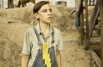 A young boy wearing a short-sleeved collared shirt, a pair of denim overalls and a large yellow lightening bolt pinned to the front of his shirt.