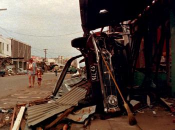 A street in Darwin that has been destroyed by Cyclone Tracy in 1974. You can see debris all over the road and a car on its side.