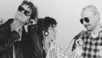 A woman in a thoughtful pose next to another woman who is holding a microphone and wearing headphones. She is standing looking at a man and pointing the microphone towards him.