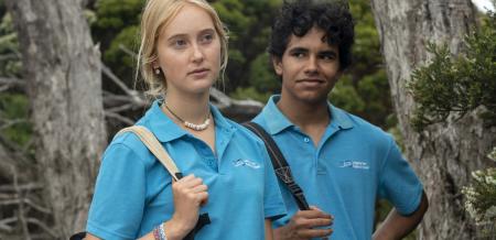 A teenage boy and girl wearing aqua shirts with a corporate logo and carrying bags. She is looking pensive, while he looks at her, smiling.