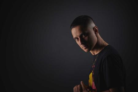 Composer DOBBY wears a gold necklace and a black t-shirt with the Aboriginal flag on it. He holds his hands in front of his chest and looks at the camera.