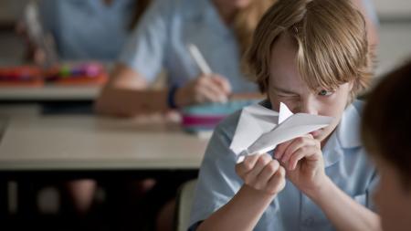 A primary school aged boy seated in a classroom focused intently on a paper plane which he is holding in his hands, ready to aim.
