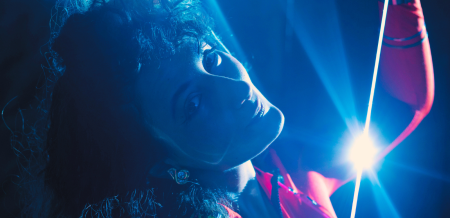 Artist Jessika Jamal Khazrik photographed under a blue light with her head turned smiling at the camera