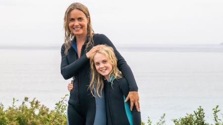 A woman wearing a wetsuit pats the head of a girl wearing a wetsuit as they walk by the ocean