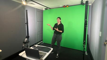 A woman presenter stands in front of a green screen and behind a small table with an open laptop on it
