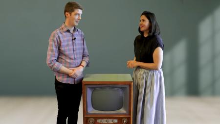 A man and woman stand looking at each other with a 1960s television set on the ground between them