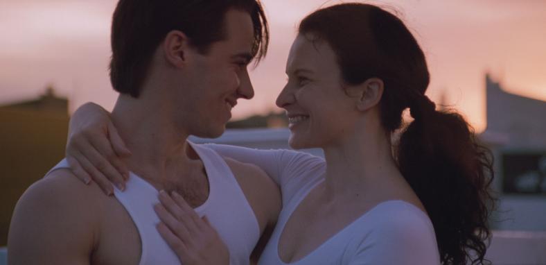 A young man and woman pictured from the shoulders up, holding each other closely and smiling at at each other. 