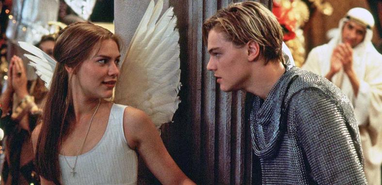 Actors Claire Danes and Leonardo DiCaprio in a scene from Romeo + Juliet. Danes is dressed in white and wearing angel wings and DiCaprio is dressed in a knight's chainmail. They are standing by a pillar at a party and looking intently at each other.