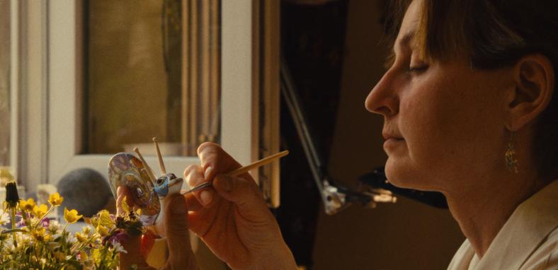 A woman handpainting a small porcelain snail while seated by an open window