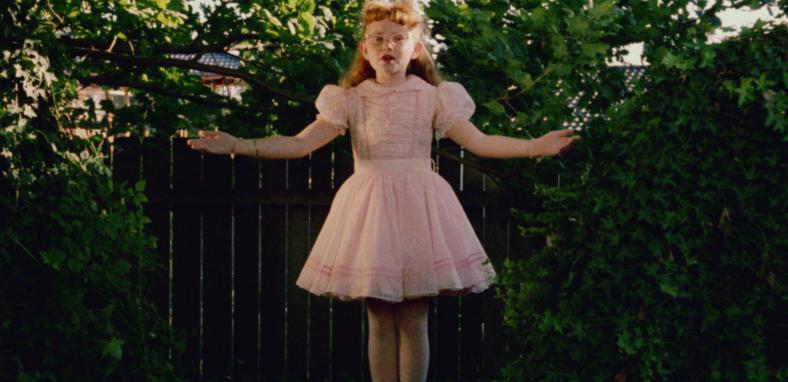 A young girl in a pink dress stands in a garden in front of a fence, with greenery behind her. Her arms are spread wide and her mouth is open.