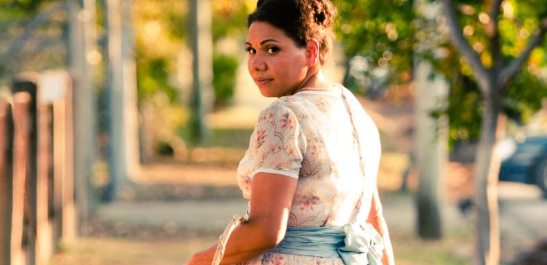 A woman in a floral dress pictured from the waist up, walking away but with her head turned to look back over her shoulder.