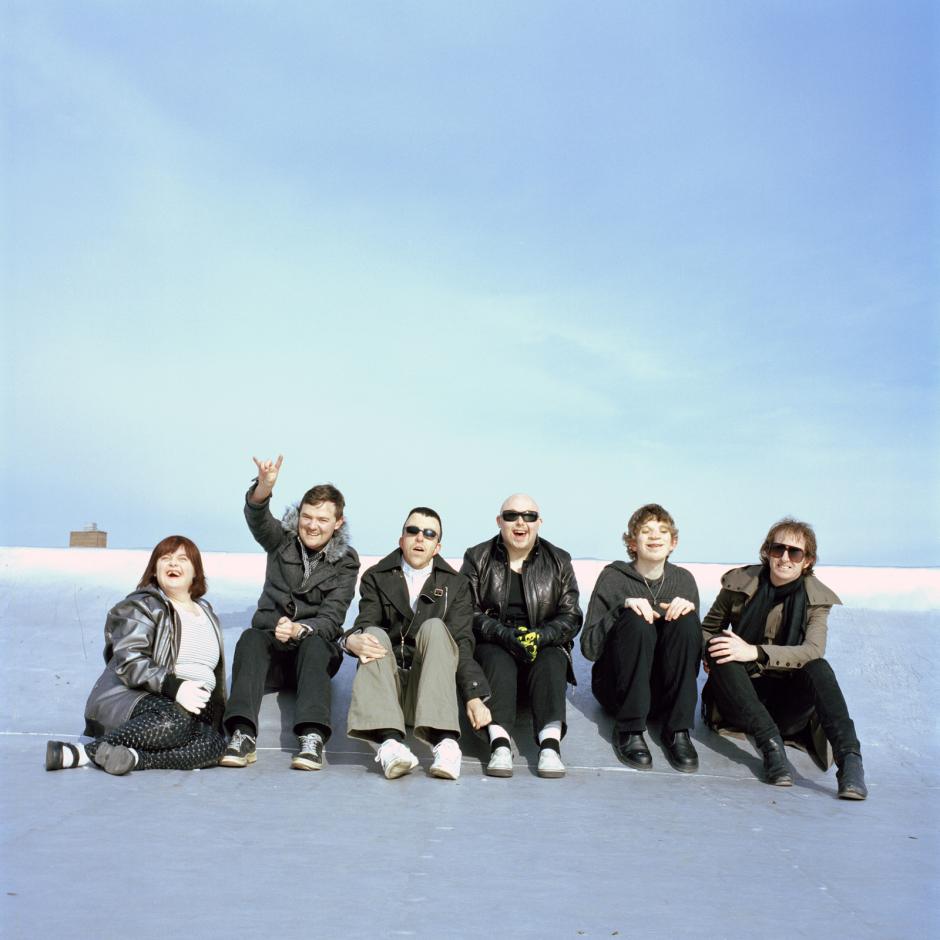 Six people sit outside on a flat surface. They are laughing and smiling and one of them is raising his hand to the sky.