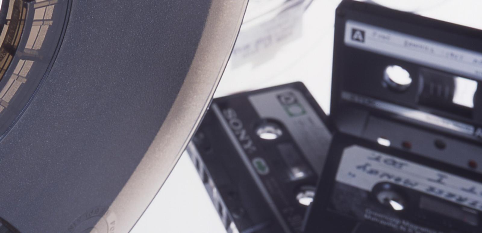 A reel to reel tape is in the foreground in front of three compact cassette tapes