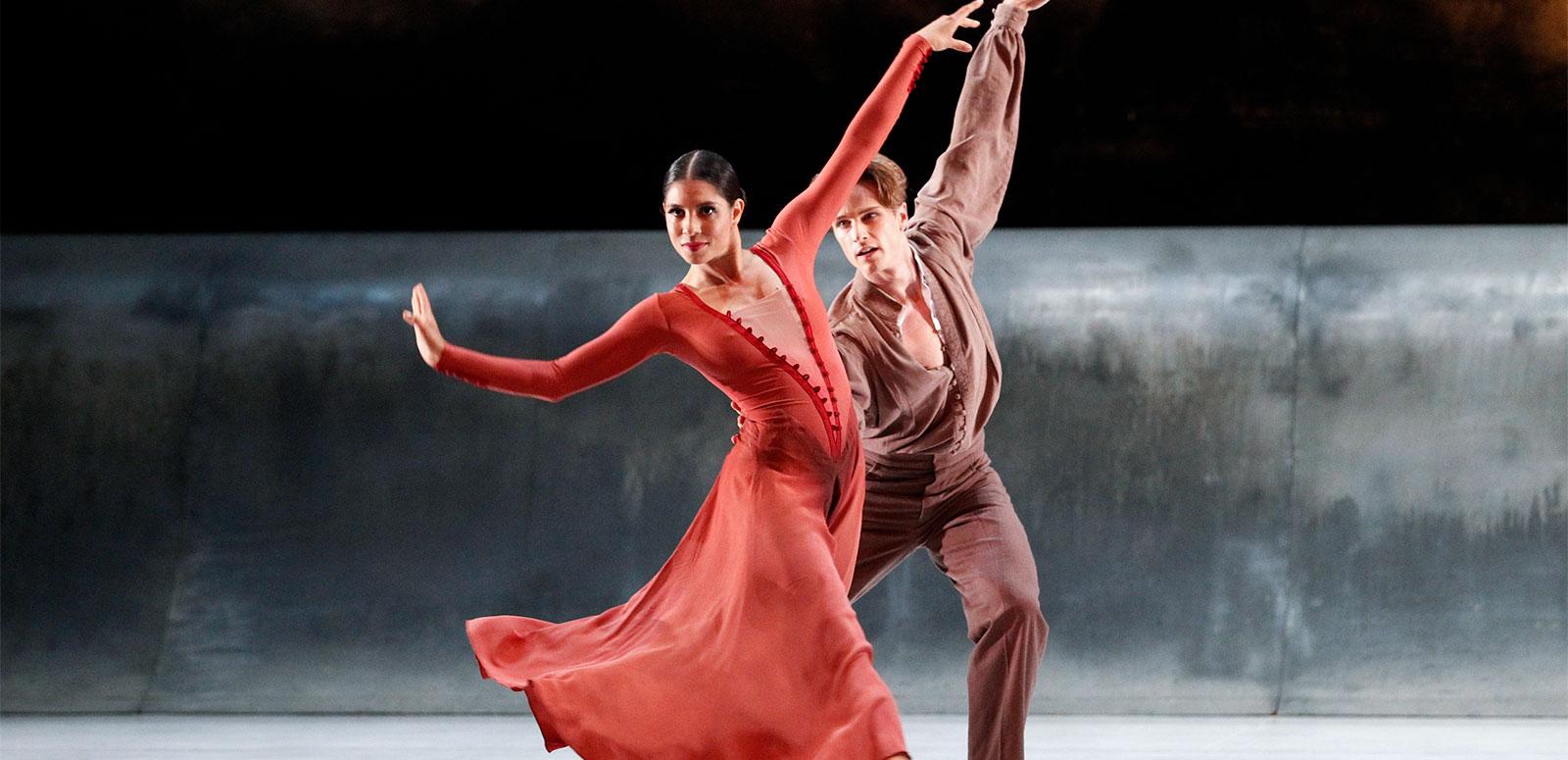 A woman dressed in red and a man dressed in brown dance on a stage against a grey and black backdrop