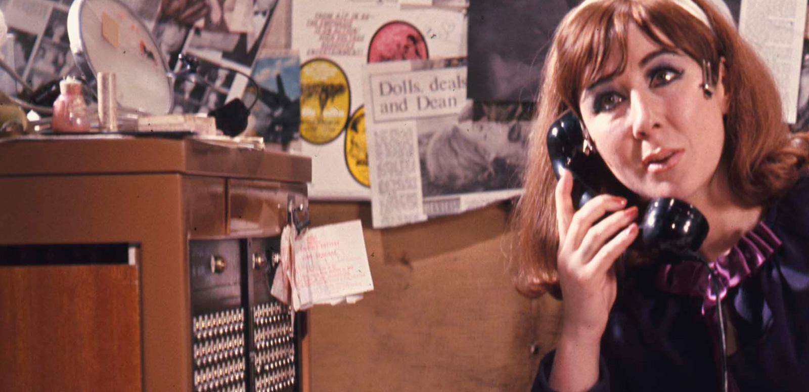 A young woman with her head tilted, holding a telephone receiver to her ear. She sits next to an old 1970s style cabinet.