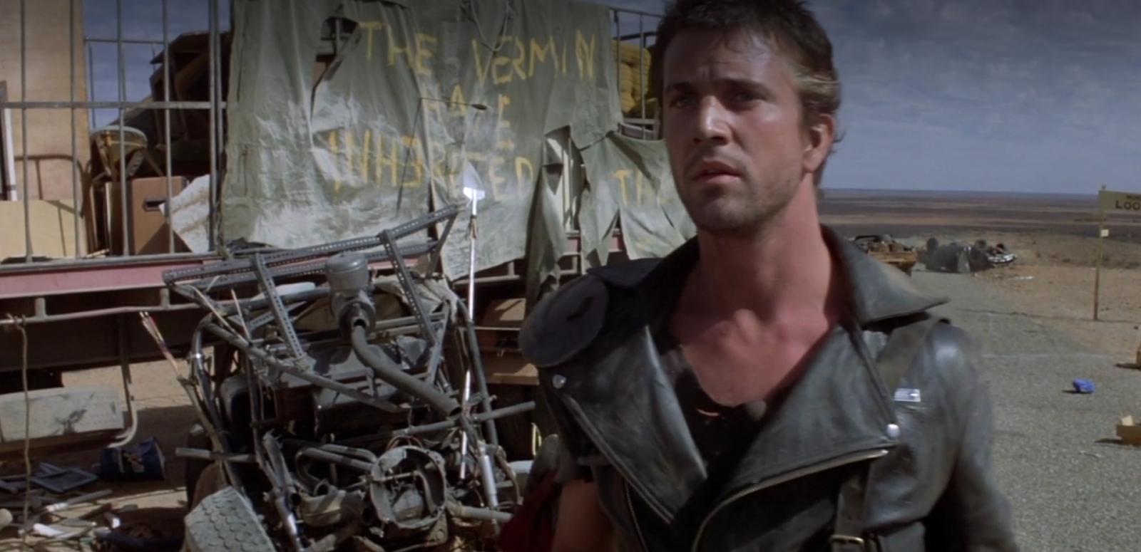 Mad Max stands next to the wreckage of a graffitied truck on a highway