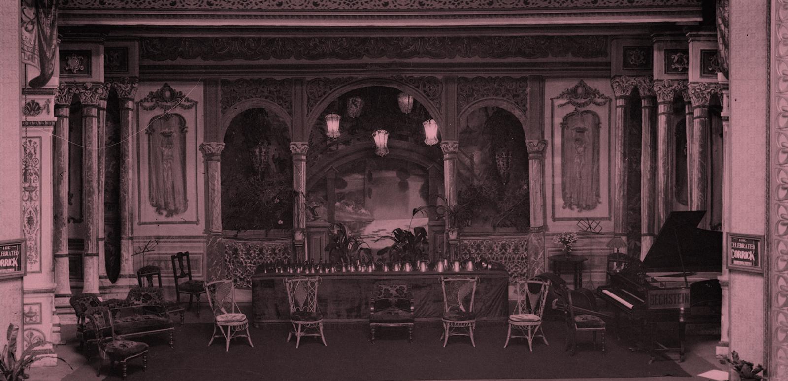 A tinted image of an early 20th century stage before a musical performance. There are a row of chairs for the performers, a piano and a table with a row of handbells.