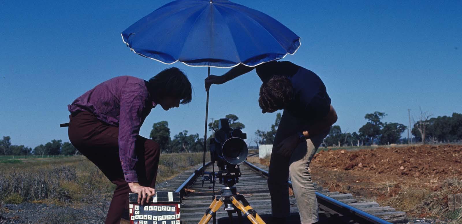 A man holds an umbrella over a film camera positioned on railway tracks while another man holds a clapperboard A man holds an umbrella over a film camera positioned on railway tracks while another man holds a clapperboard