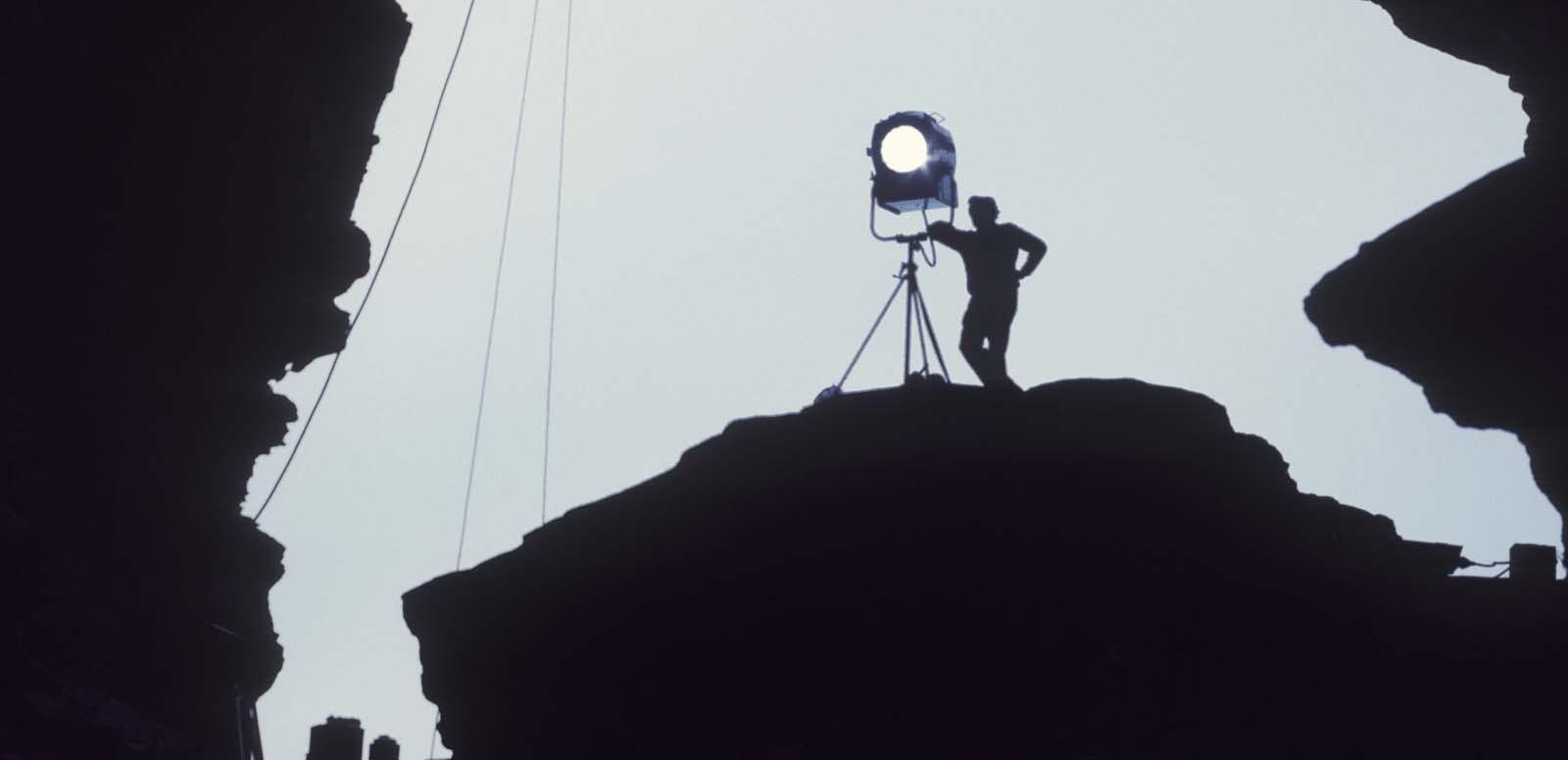 A member of a film crew stands silhouetted next to a standing light on an outcrop of rock