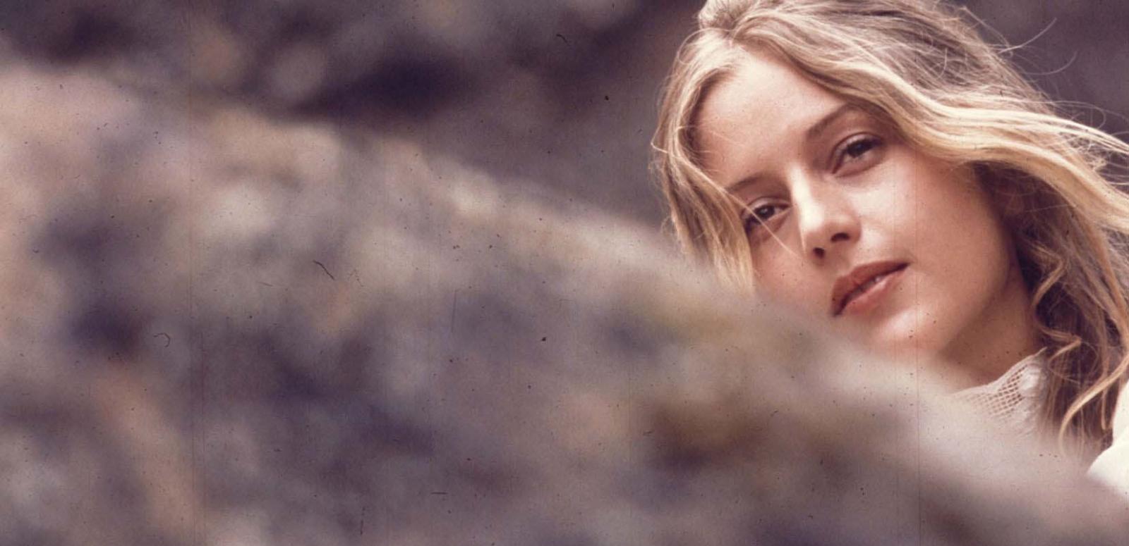 Film still from Picnic at Hanging Rock with Anne Lambert as Miranda looking winsome with her face partially obscured by a boulder in the foreground.