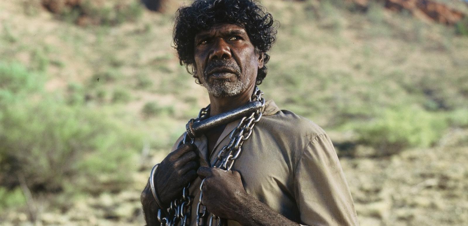 An aboriginal man stands chained in an arid landscape.