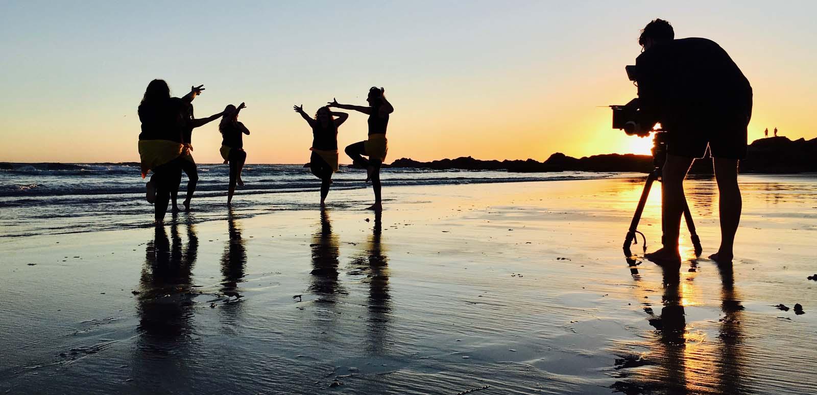 A camera filming silhouetted Aboriginal dancers on a Byron Bay beach at sunrise