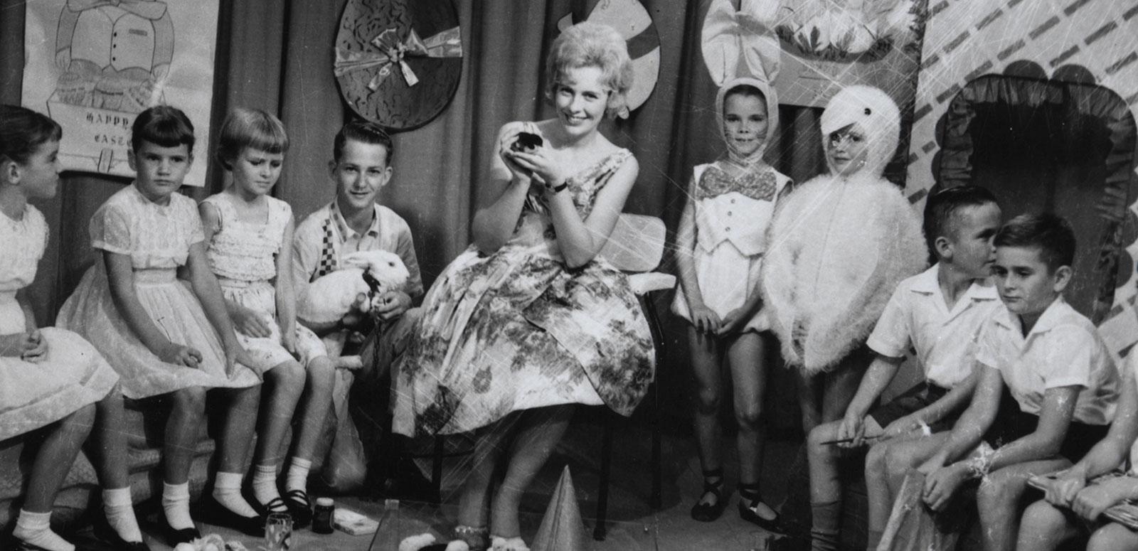 A smiling woman seated in a TV studio surrounded by children. 