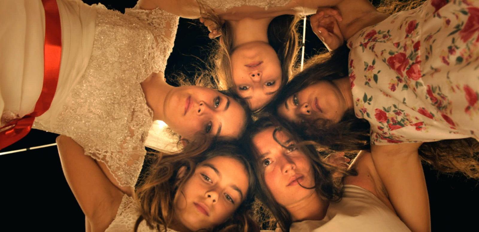 5 young women leaning over linking arms in a circle and looking directly down into the camera.