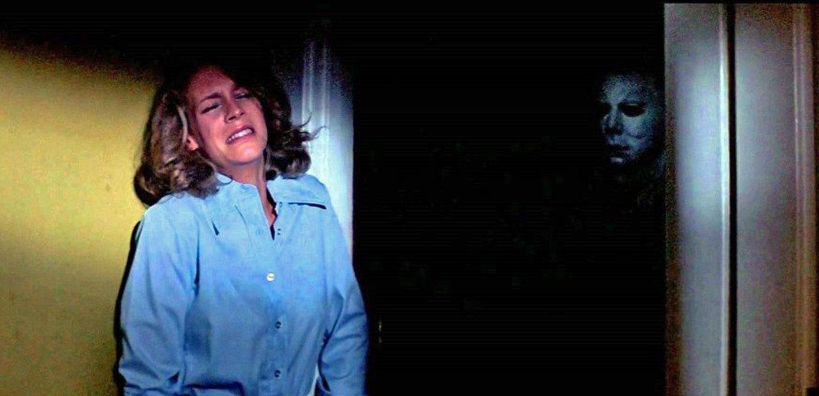 A terrified woman standing against a wall while a shadowy figure in a white mask stands behind her.