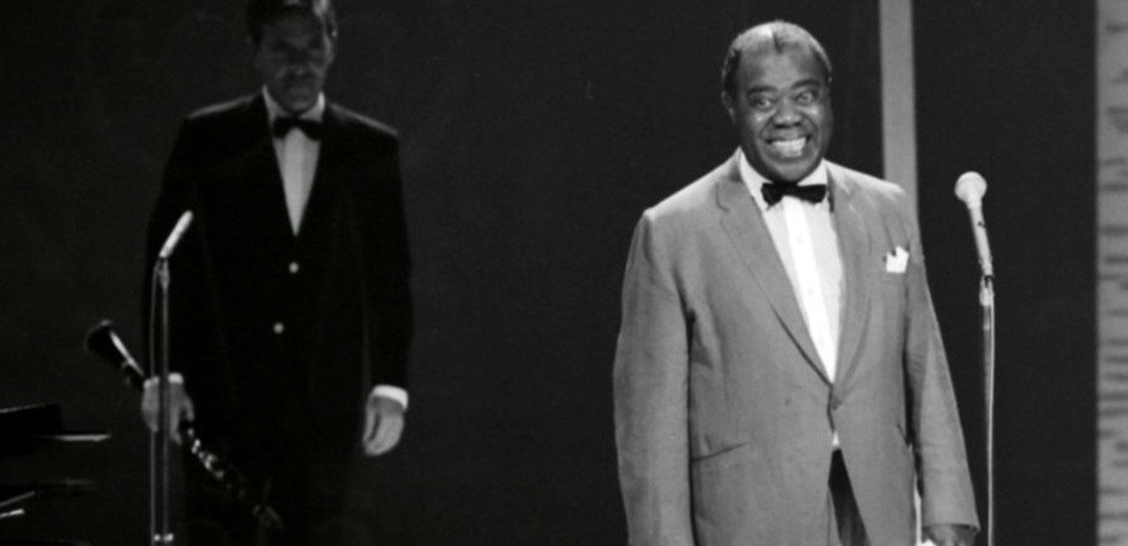 Jazz musician and singer Louis Armstrong standing in front of a microphone in a TV studio, smiling. Another musician is pictured beside him holding an instrument. Circa 1964.