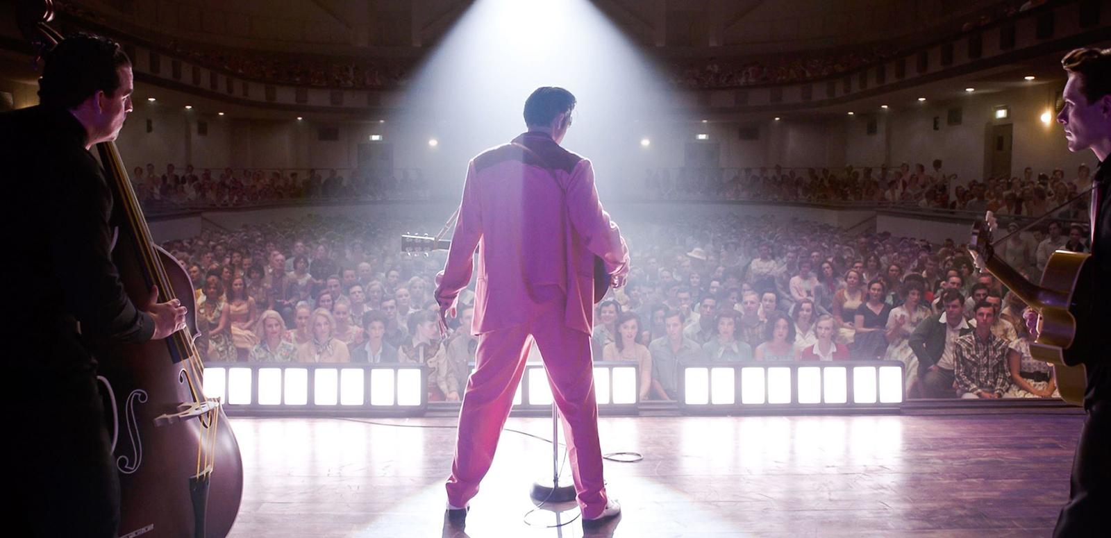 Actor Austin Butler in a scene from the film ELVIS. He is pictured from behind wearing a bright pink suit and standing on a stage in front of a large crowd in a theatre.