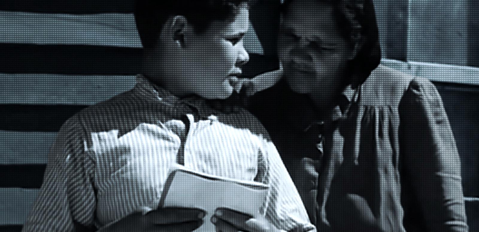 A schoolboy holding a notepad while someone looks over his shoulder at it.