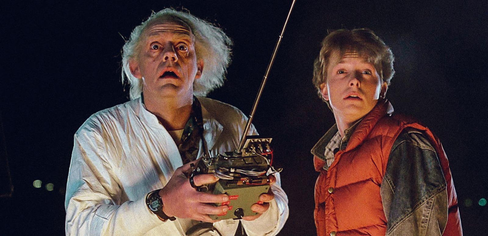 An older man with white hair, wearing a lab coat and holding a large remote control standing next to a young man at night time, they both look shocked as they stare into the distance.