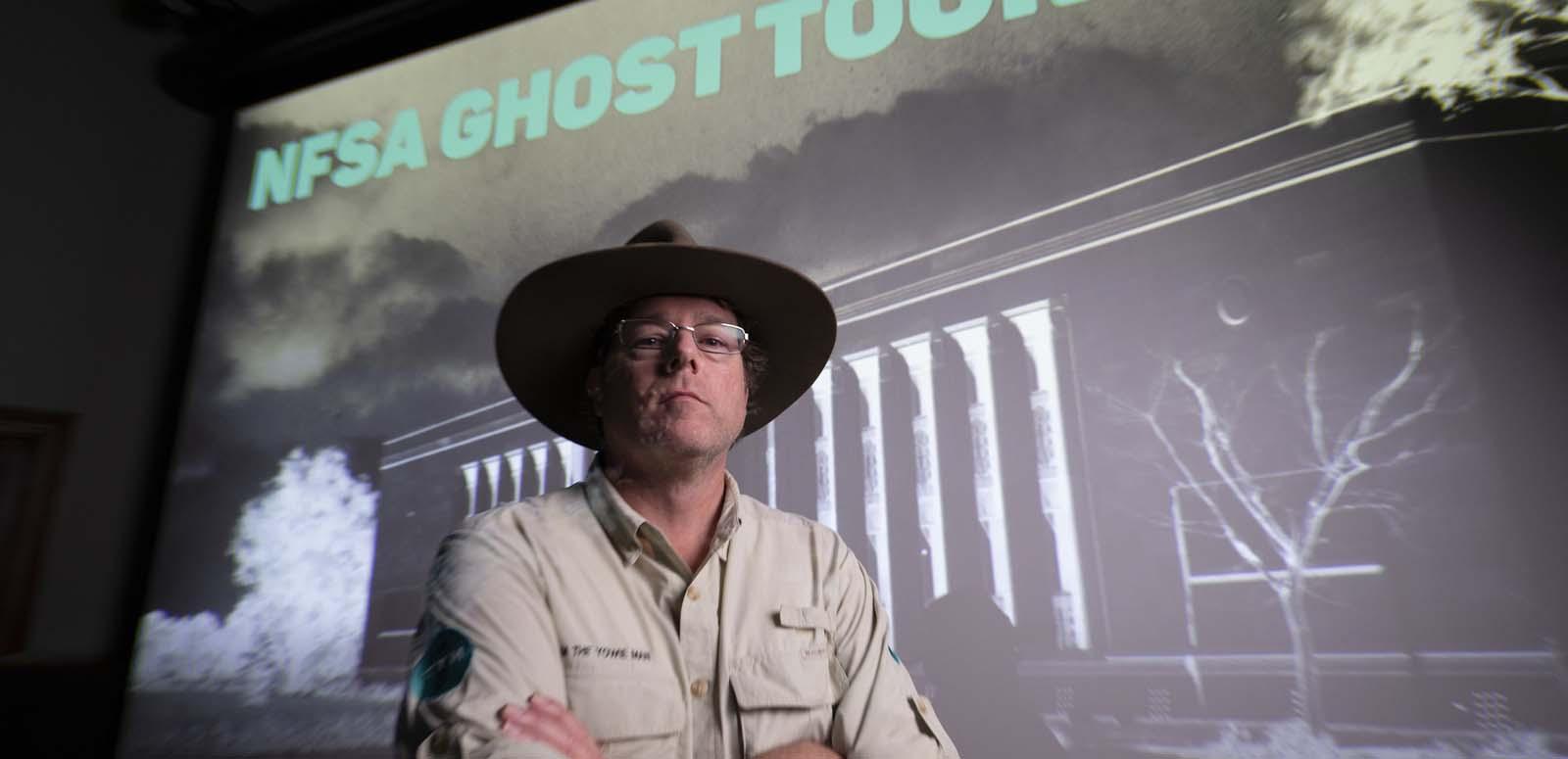 Tim the Yowie Man in the NFSA Theatrette standing before a slide of the NFSA during an evening Ghost Tour