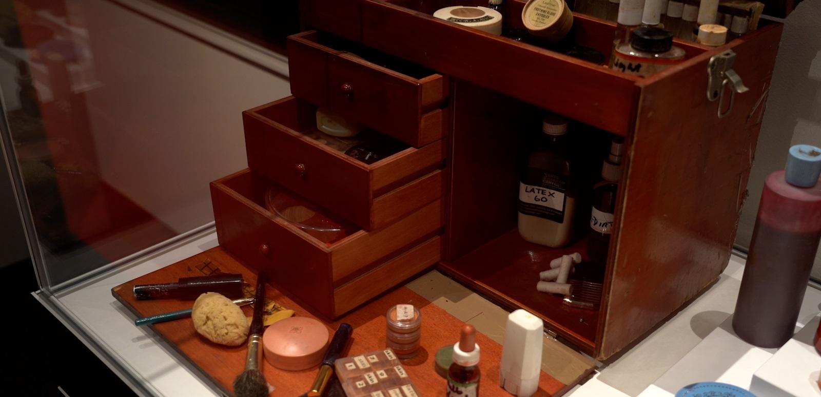 Wooden make-up chest belonging to film make-up artist Vivien Mepham. The chest's drawers are open and make-up jars, brushes and bottles are displayed in front of the chest