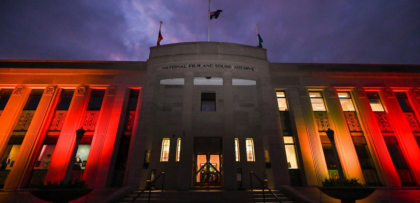 Front facade of an art deco style building with columns at the front and a sign saying National Film and Sound Archive. The building is lit up with red and orange lighting at night time.