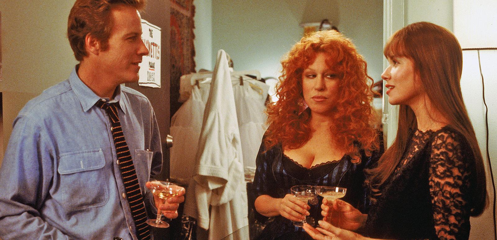 A man and two woman, roughly in their 30s, stand holding glasses of wine talking to each other in a scene from the film Beaches. One of the women looks unhappy.