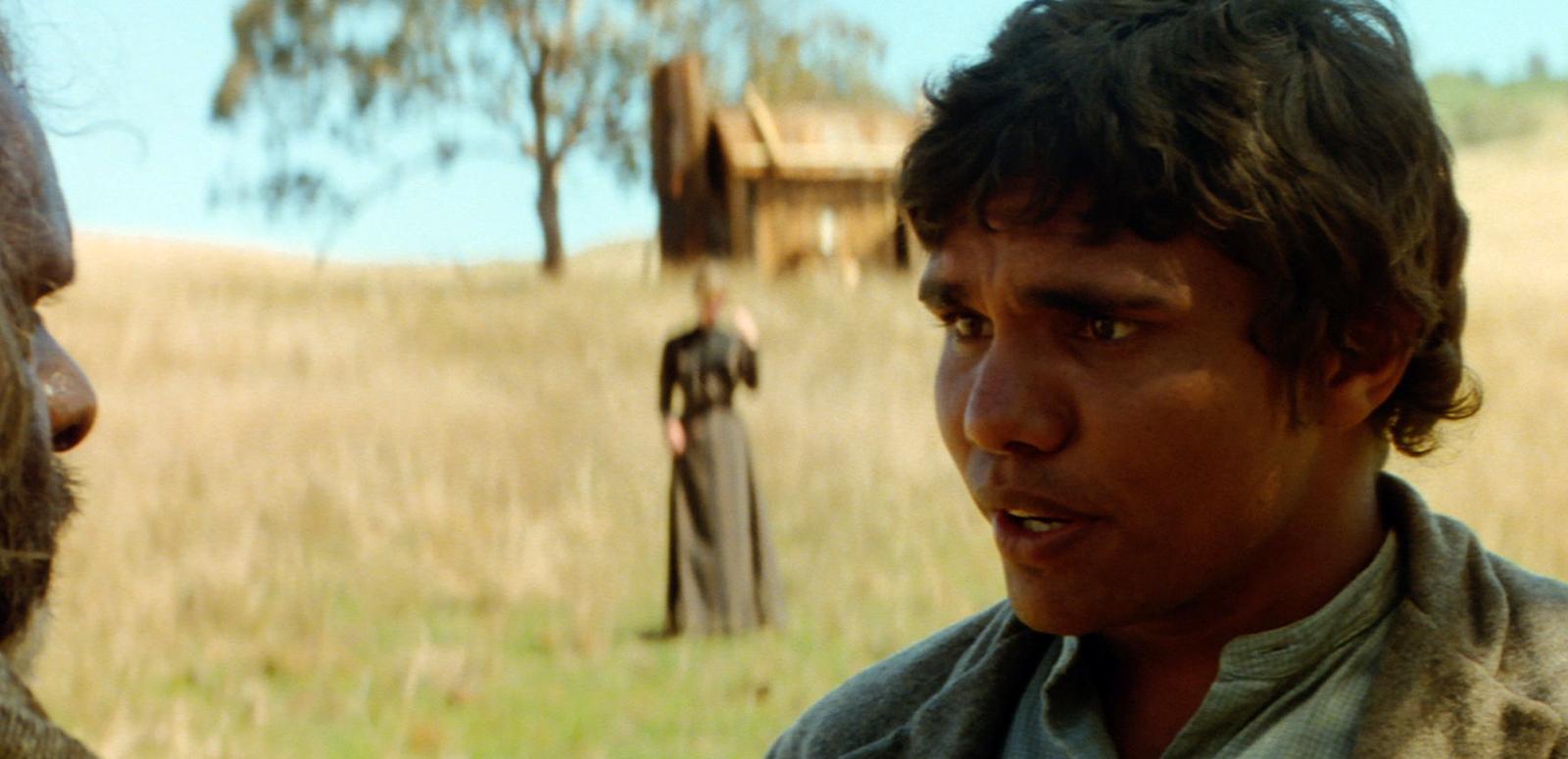 Close up of a young Indigenous man talking to an older man whose face is just visible on the left. A woman is in the distance and they appear to be in an outback setting.