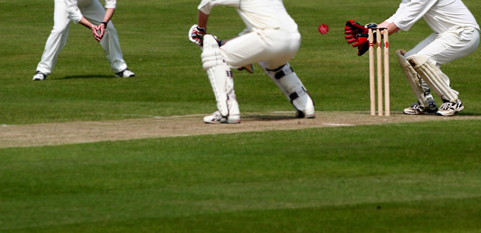 Close up on cricket pitch with focus on lower body shots of batsman, wicket-keeper and fielder engaged in play.