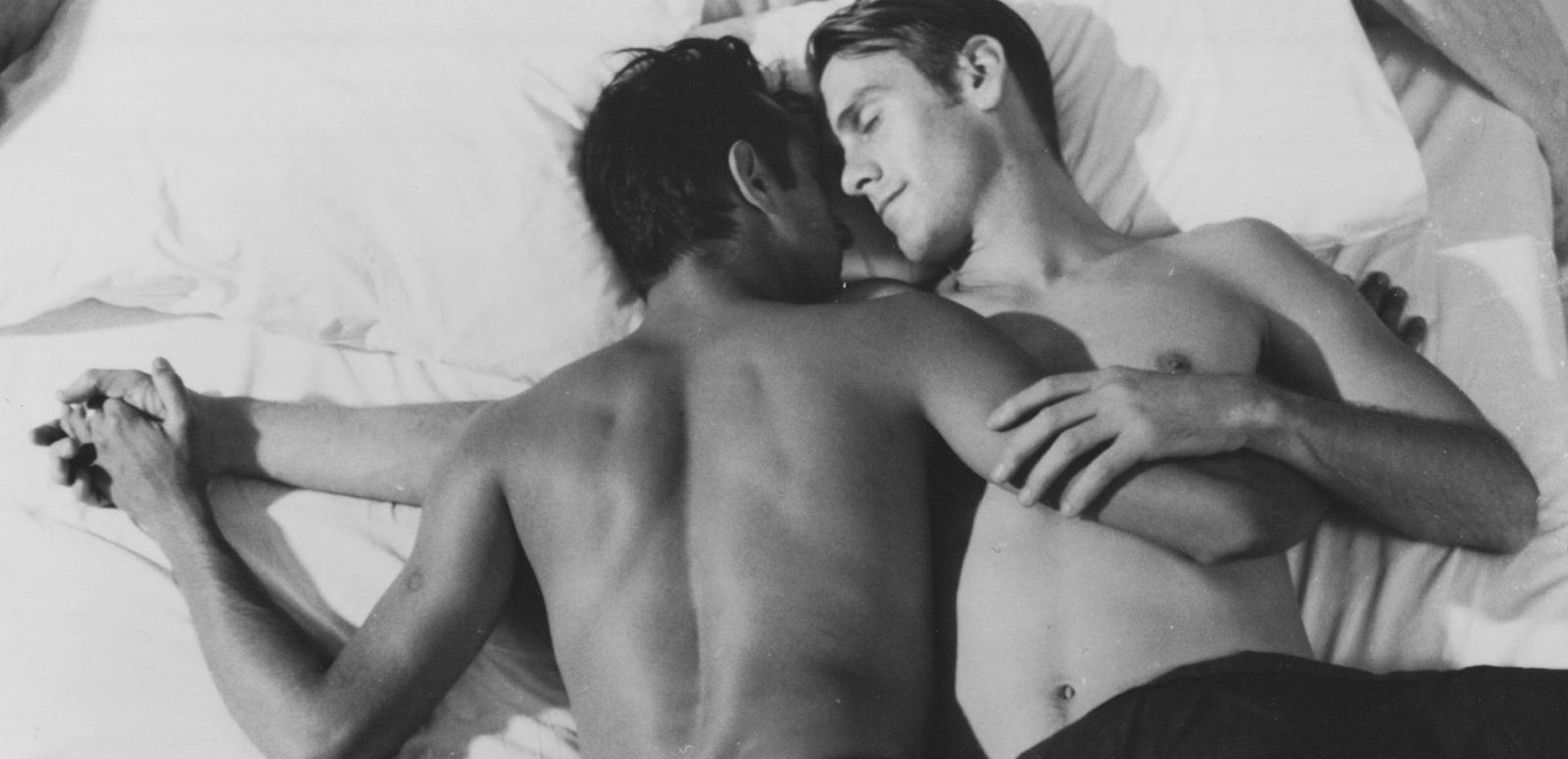 Two young men lying on a bed in an embrace. They are both shirtless and one is lying face down and the other facing up, with their faces turned toward eachother.